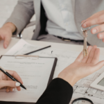 Entrega de chaves entre duas pessoas durante assinatura de contrato imobiliário, com planta arquitetônica e tablet sobre a mesa.