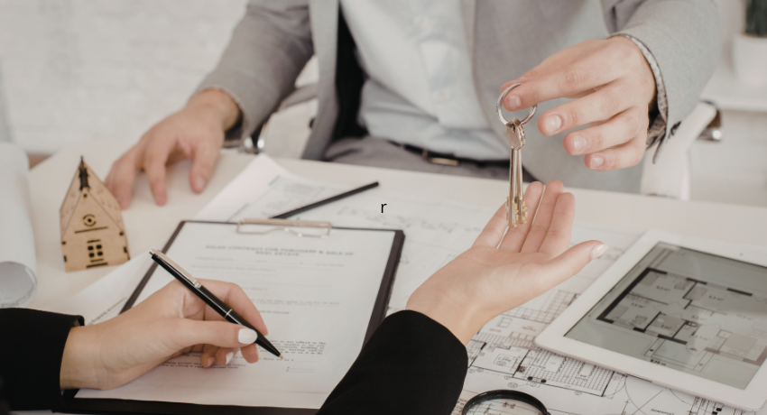 Entrega de chaves entre duas pessoas durante assinatura de contrato imobiliário, com planta arquitetônica e tablet sobre a mesa.